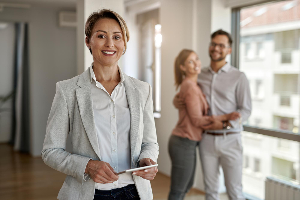 Professional real estate agent standing confidently in a modern apartment with a happy couple in the background.